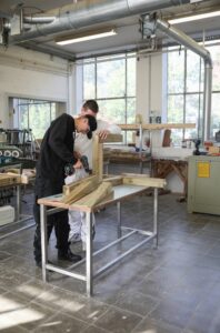 Ein junger Mann arbeitet mit einem Akkuschrauber in einer Werkstatt. Dahinter steht eine weitere Person und schaut zu.